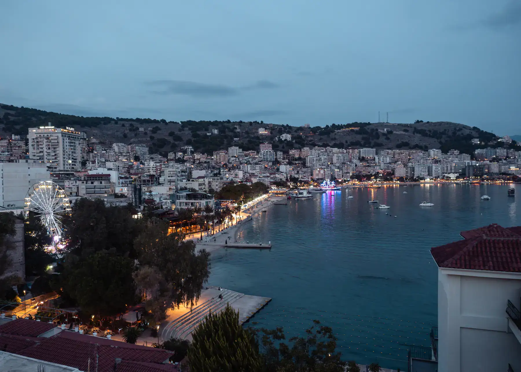 View of Saranda, white apartment building along the waterfront, with mountains in the background, at dusk with lights along the promenade.