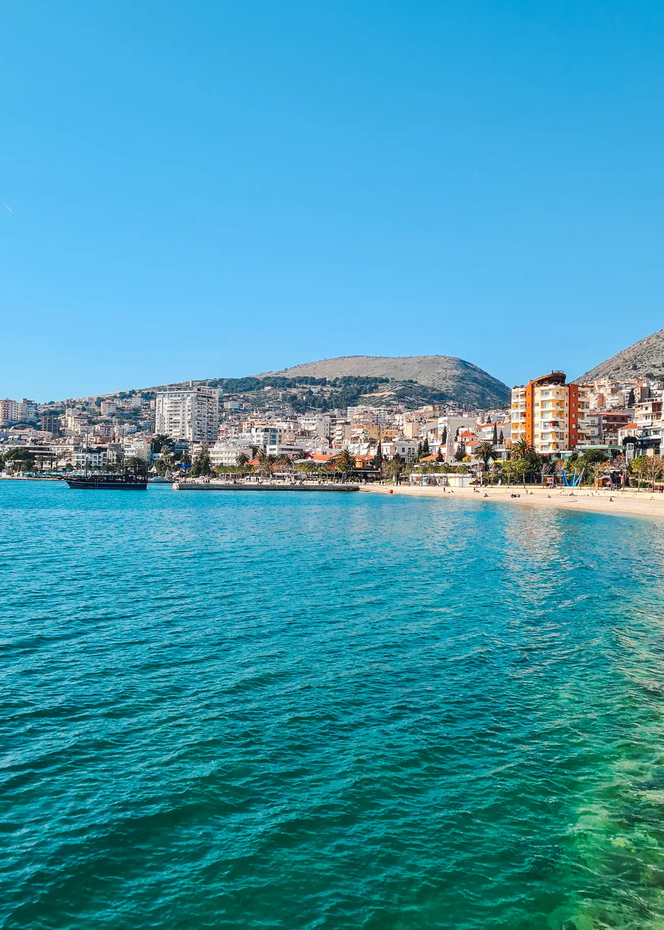 View of the turquoise ocean and one of Saran's public beaches with high-rise apartment buildings in the background on a sunny day in Sarand&euml; Albania.