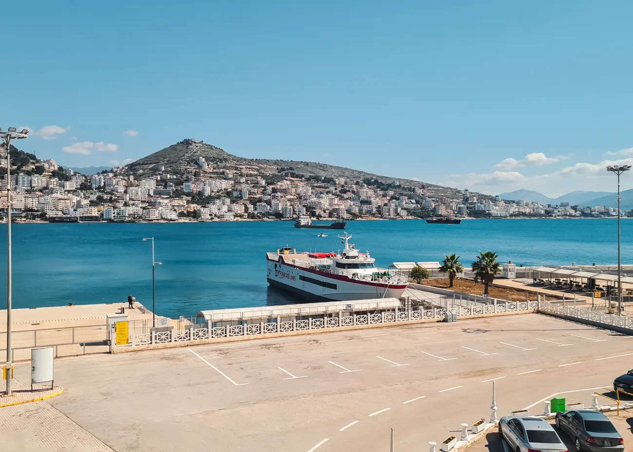 View of a Finikas Lines red and white ferry docked at Saranda Ferry Port with Saranda town and mountains in the background on a sunny day,