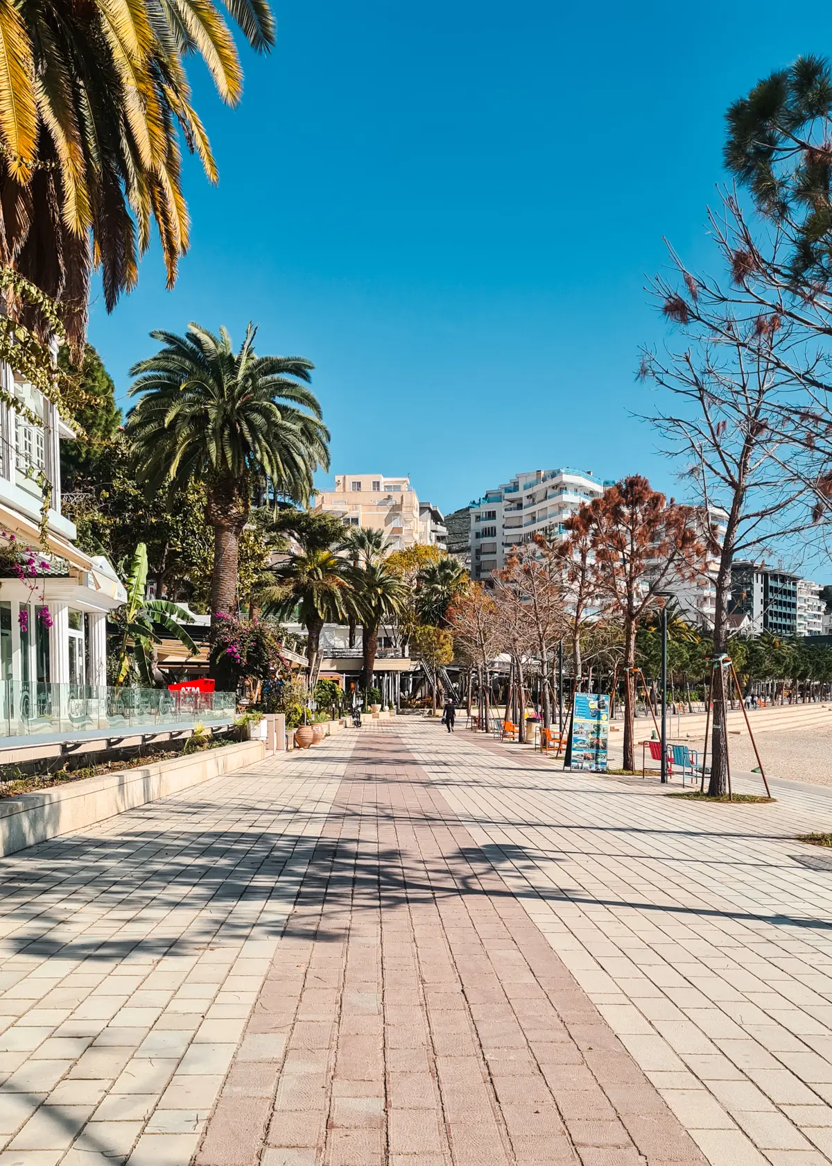 Paves Hasan Tahsini Boulevard (the promenade in Saranda), lined by benches, trees and the public beach on the right, and palm trees and shops on the left.
