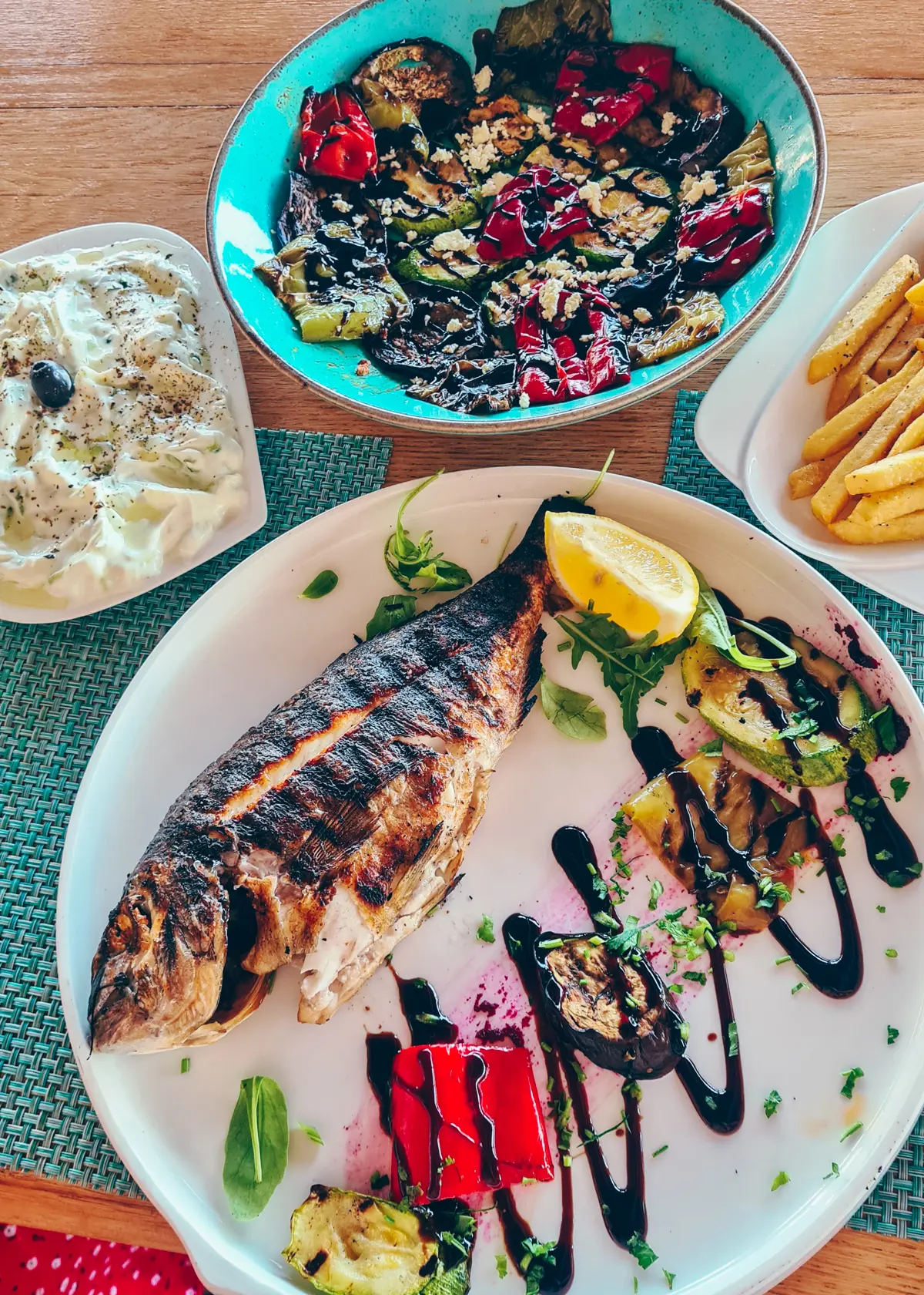 Large white plate with a whole grilled fish and some garnish, blue plate with grilled vegetables, white dish with tzatziki and fries on a table in Summer Dream Restaurant at Drymades Beach on the Albanian Riviera.