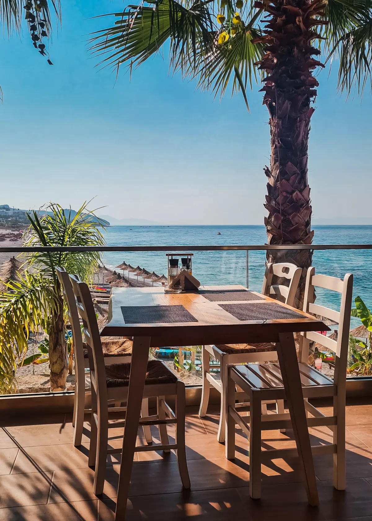 Wooden table and four chairs by a glass railing with a large palm tree on the balcony of Summer Dream Restaurant at Drymades Beach.