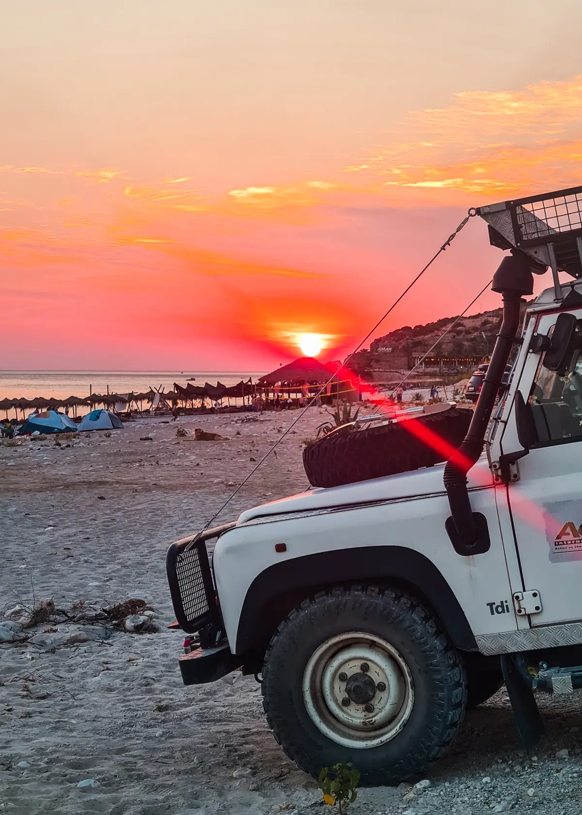 Close up of a white old Jeep on Drymades Beach with a orange, yellow and red sunset in the background.
