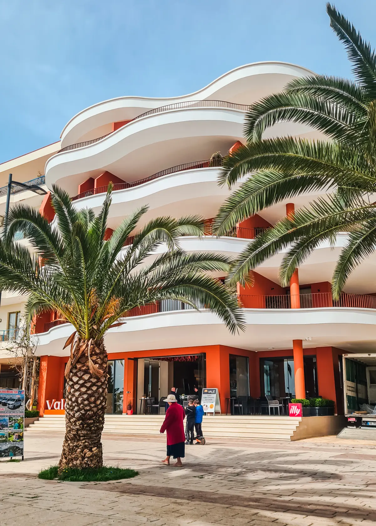 Orange and white hotel withe Vale Hotel, with organic shaped verandas and two palm trees in front in Saranda, Albania.