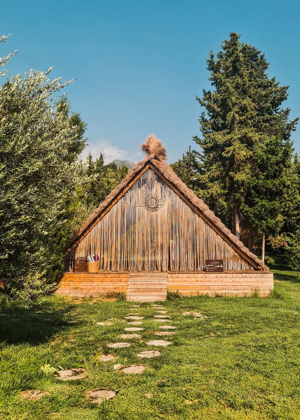 A-shaped Moyo Yoga Shala Ade from wood and straw set in a lush garden with a stone pathway leading to it on a sunny day at Drymades Beach.