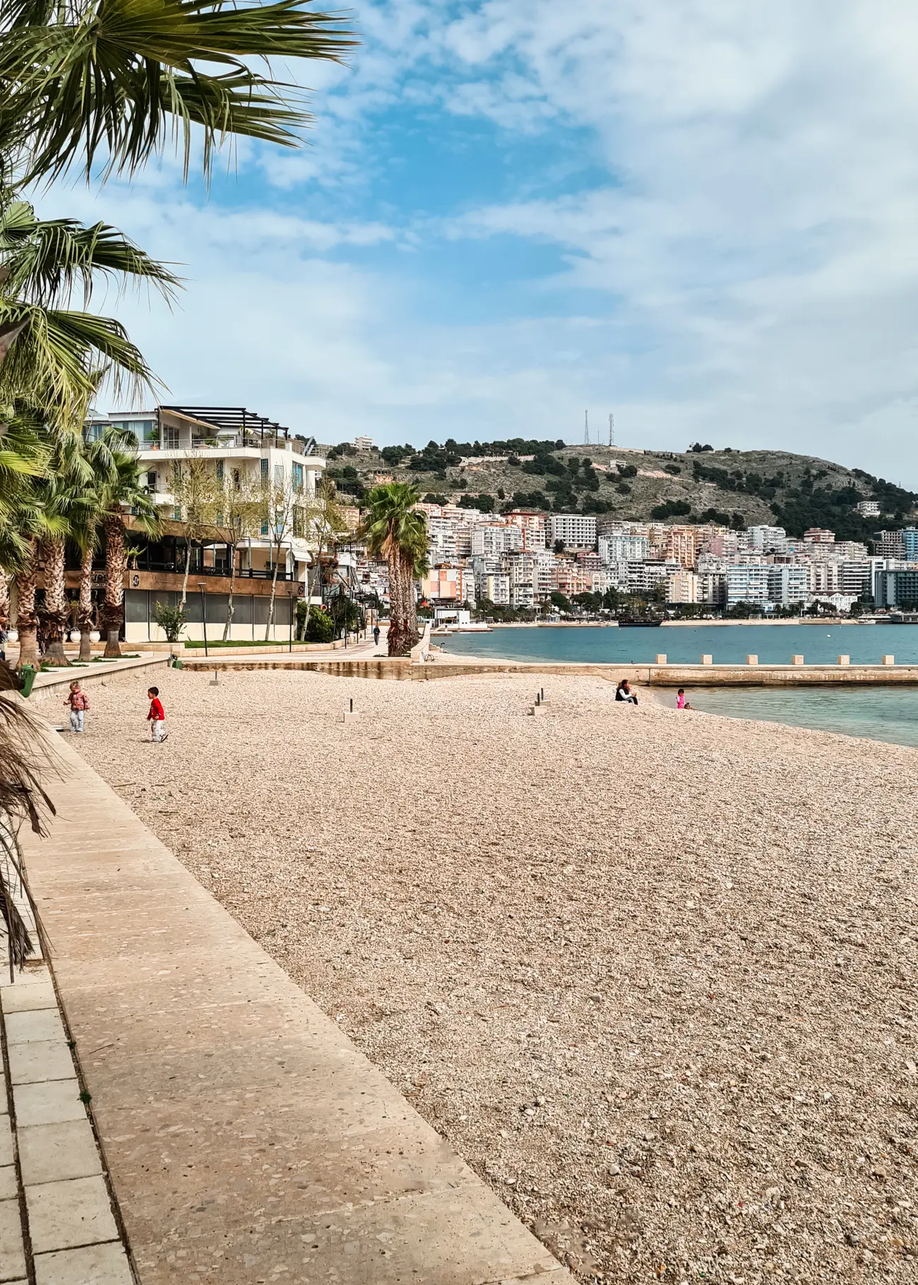 A pebbled public beach in Saranda, lined by palm trees with the city in the background.
