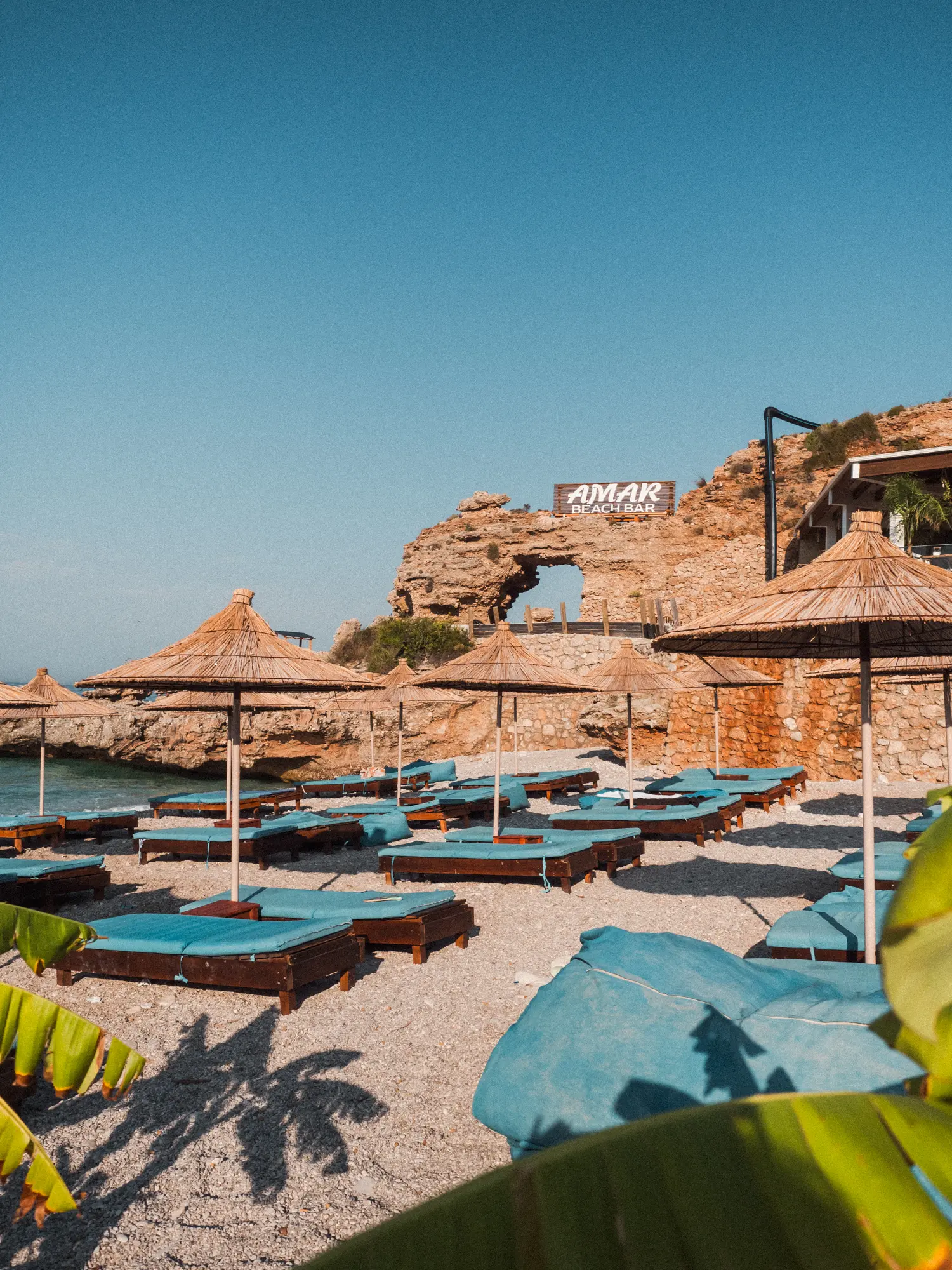 Blue sunbeds, straw parasols on a pebble beach with a stone arch in the background on Drymades Beach close to Dhermi in Albania.