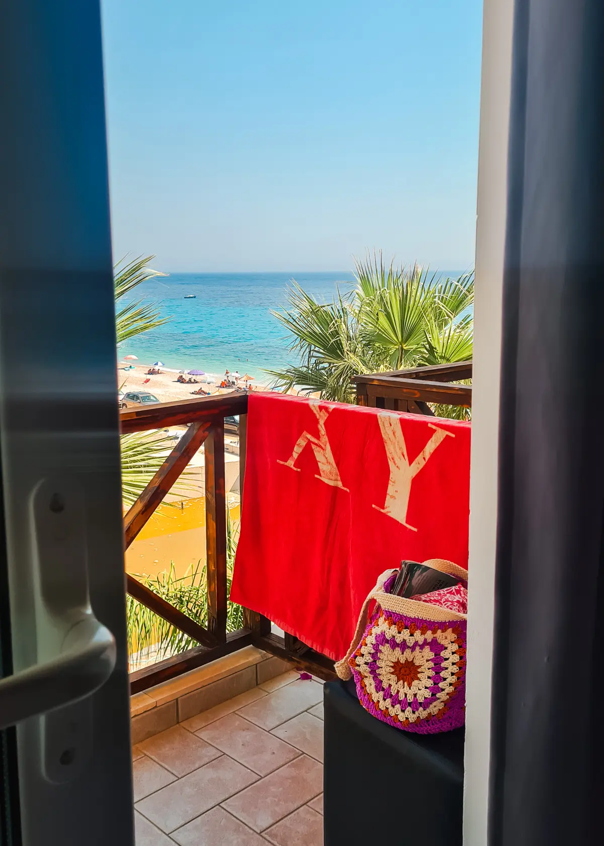 White and purple crochet bag and a red Roxy beach towel on a balcony at Summer Dream Hotel overlooking Drymades Beach and turquoise ocean on the Albanian Riviera.