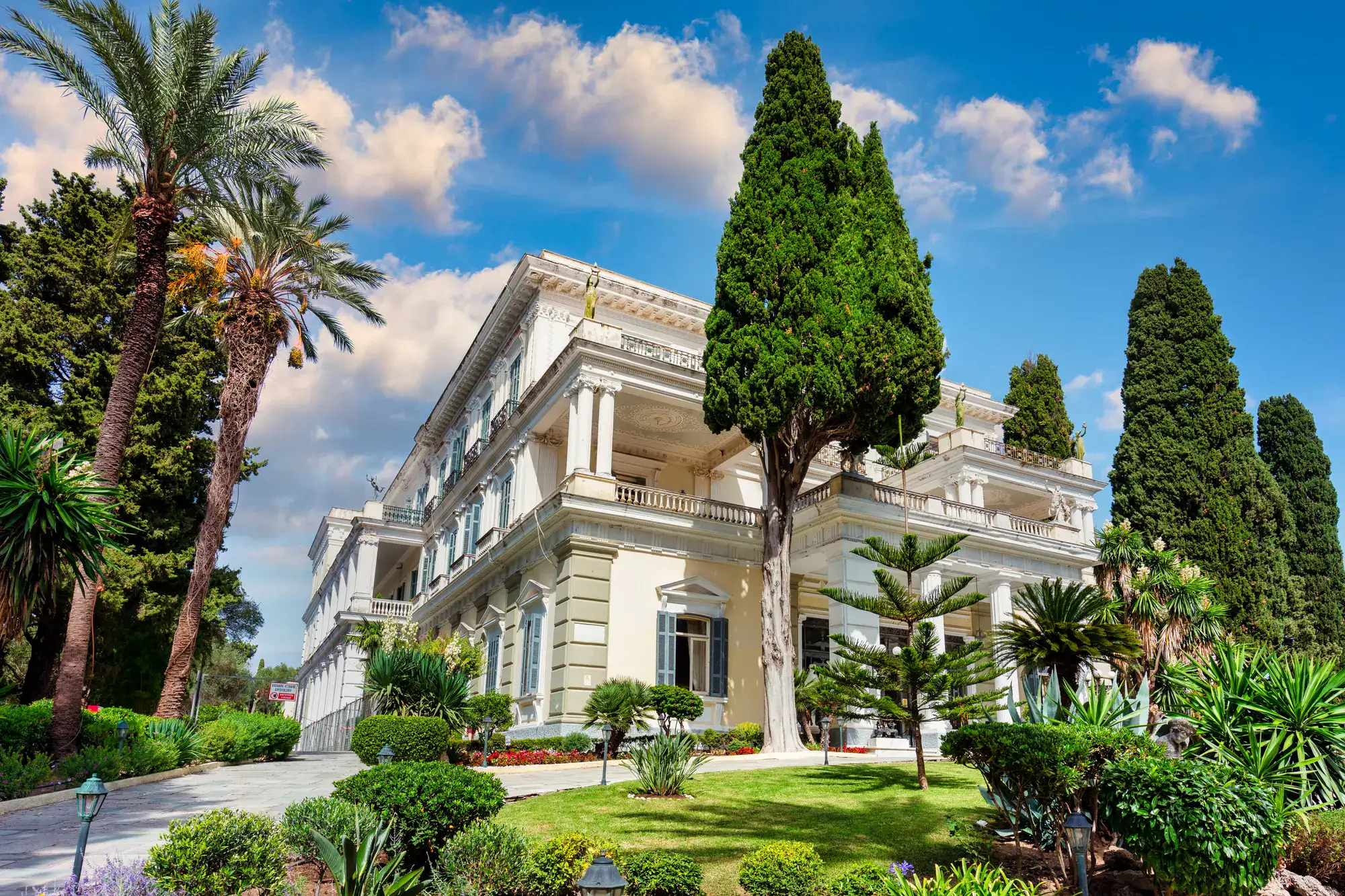 Majestic Neo-Classical Achilleion Palace with a tall cone shaped green tree and green grass in the front, palm trees on the left, during a one day Corfu itinerary.