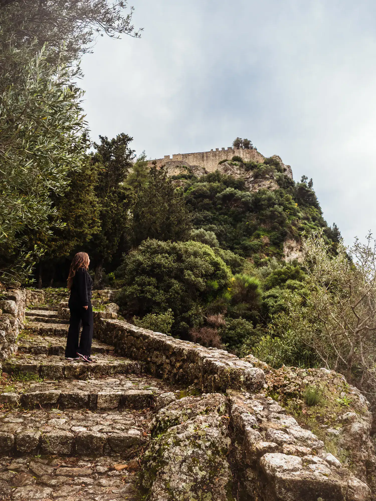 Woman wearing a black sweater and pants, staring on an old stone staircase leading through the forest to Angelokastro Fortress during a one day itinerary in Corfu.
