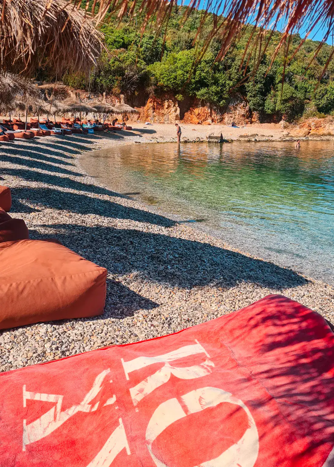 Red towel with white letters, orange bean bags in the background along the shore of the pebbly  Augustus Beach in Ksamil.