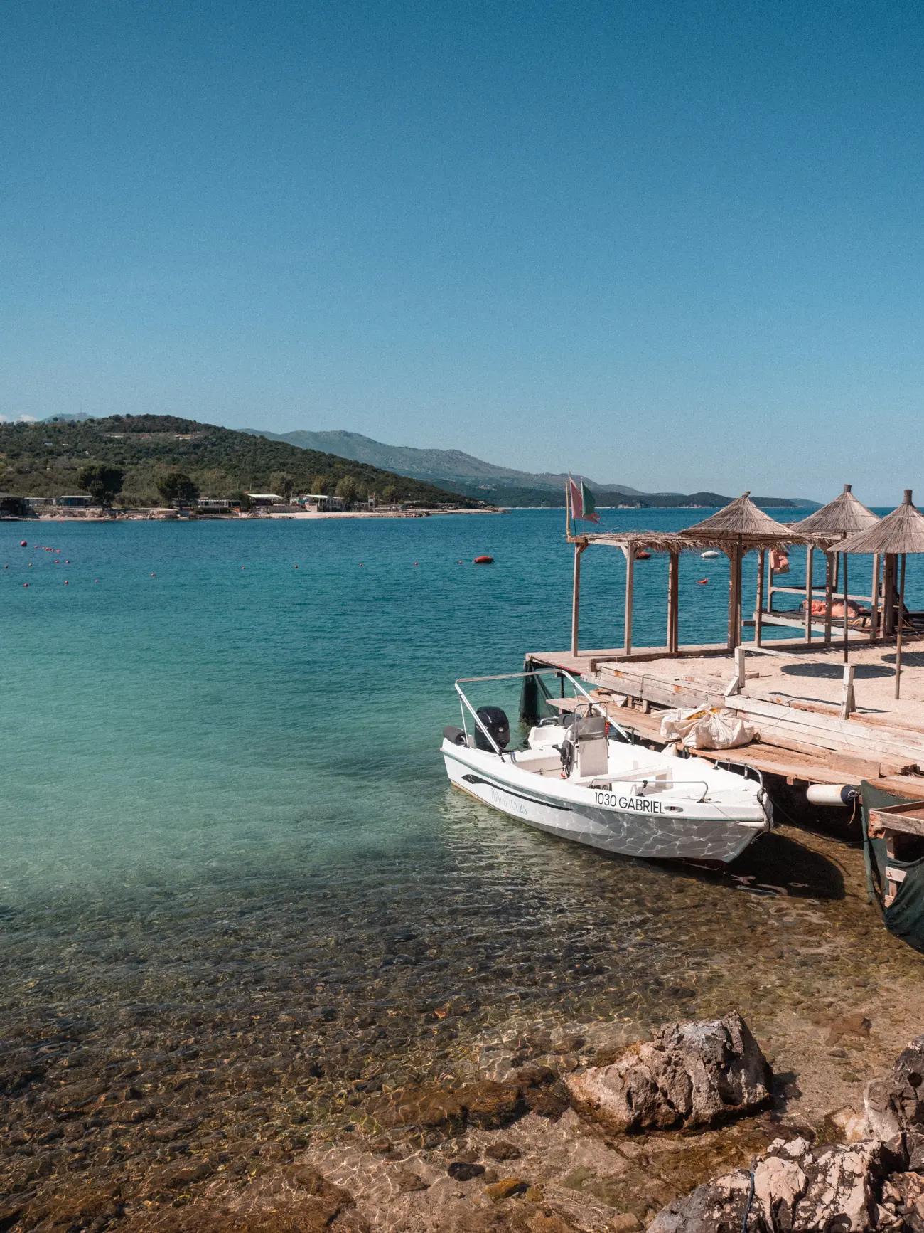Small white boat docked next to a wooden deck with parasols above the turquoise ocean with a green island in the background, one of the best beaches is Ksamil.