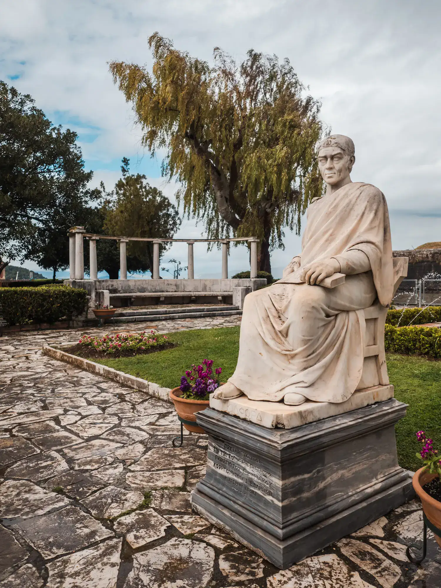 White statue of a man sitting on a chair in roman clothes, with a rounded bench with pillars in the background on a gloomy day in Boschetto Garden, Corfu Old Town.