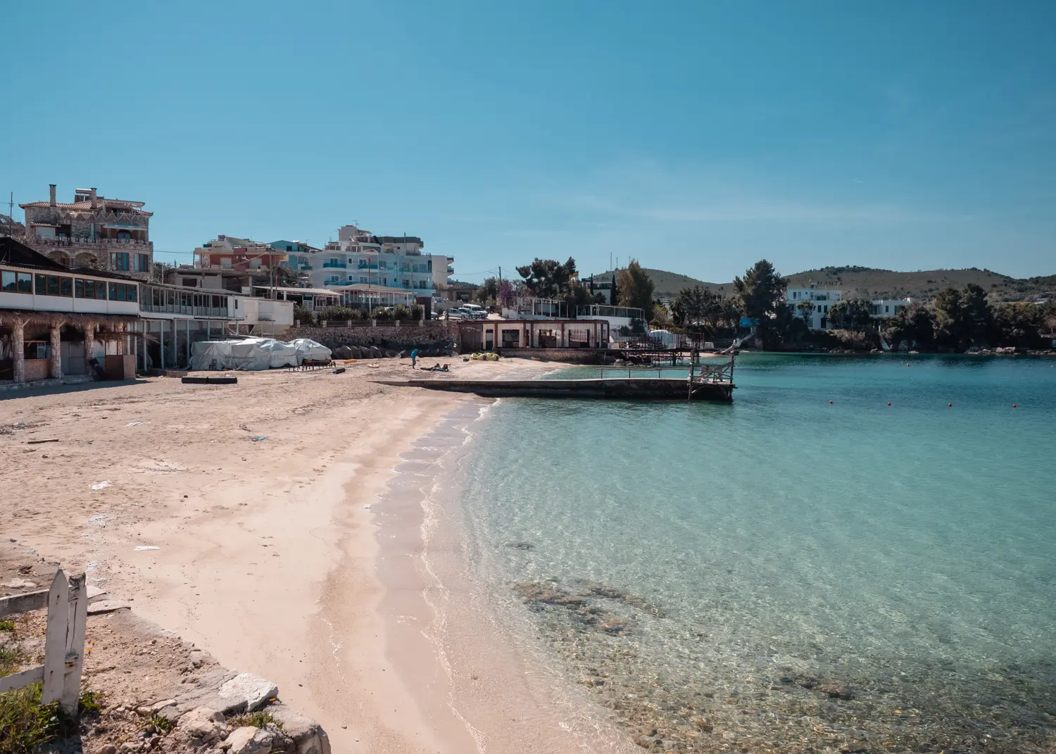 Light colored sandy beach with a concrete jetty in the middle, hotels in the background and shallow light blue water, Ksamil Beach guide.