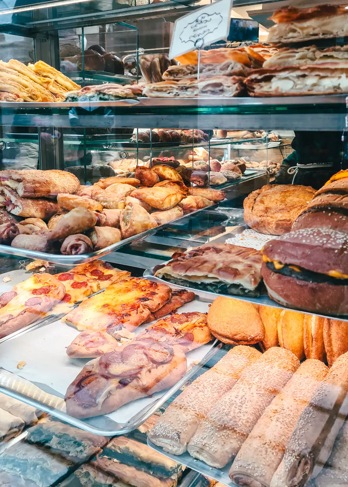 View from outside of a glass counter filled with pastries, pizza, burgers at Panetteria, the best bakery in Corfu Old Town.