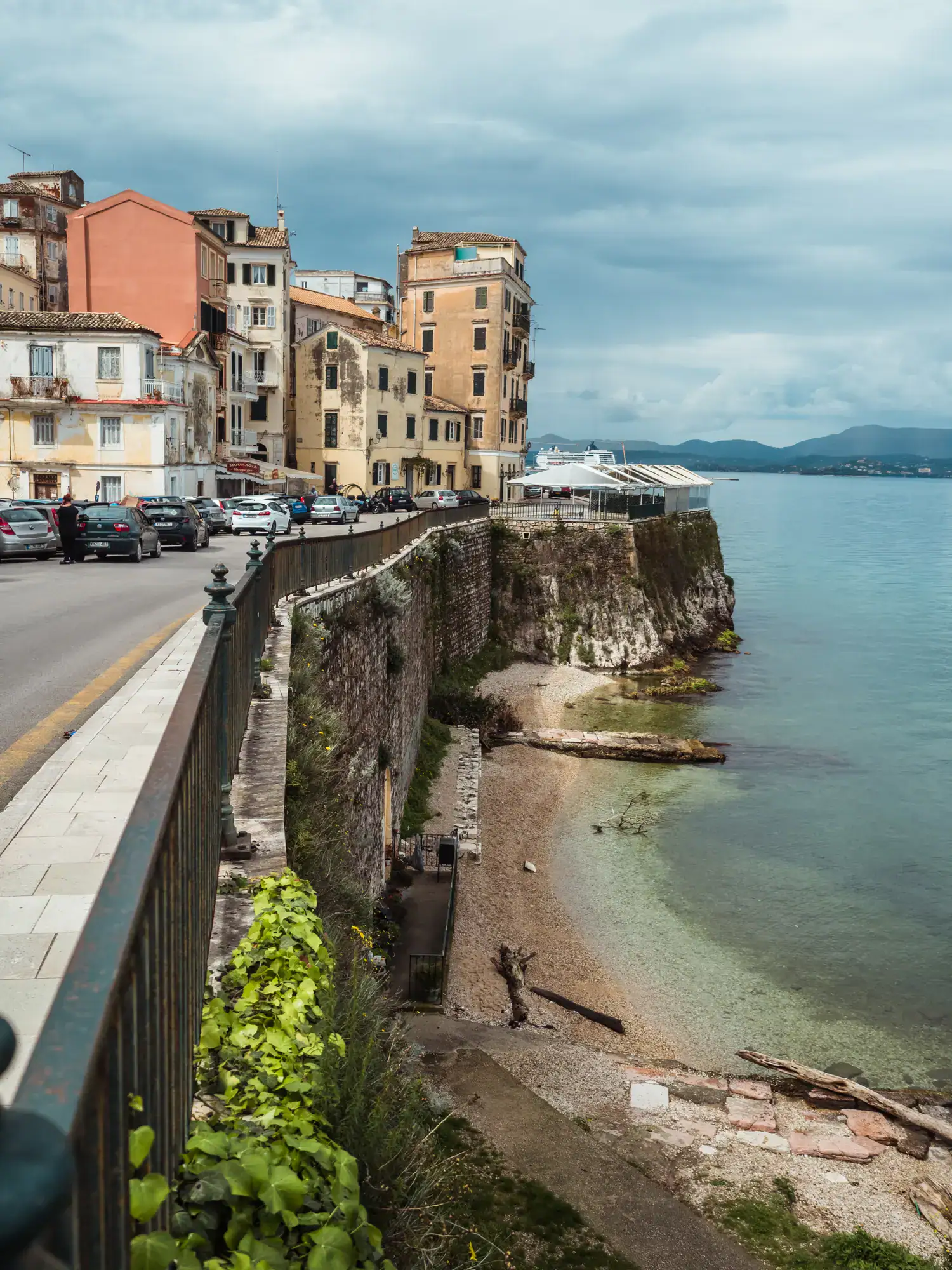 Narrow pebbled beach along the old wall of Corfu Town, with white, yellow and peach apartment buildings in the background on a gloomy day exploring Corfu Old Town.