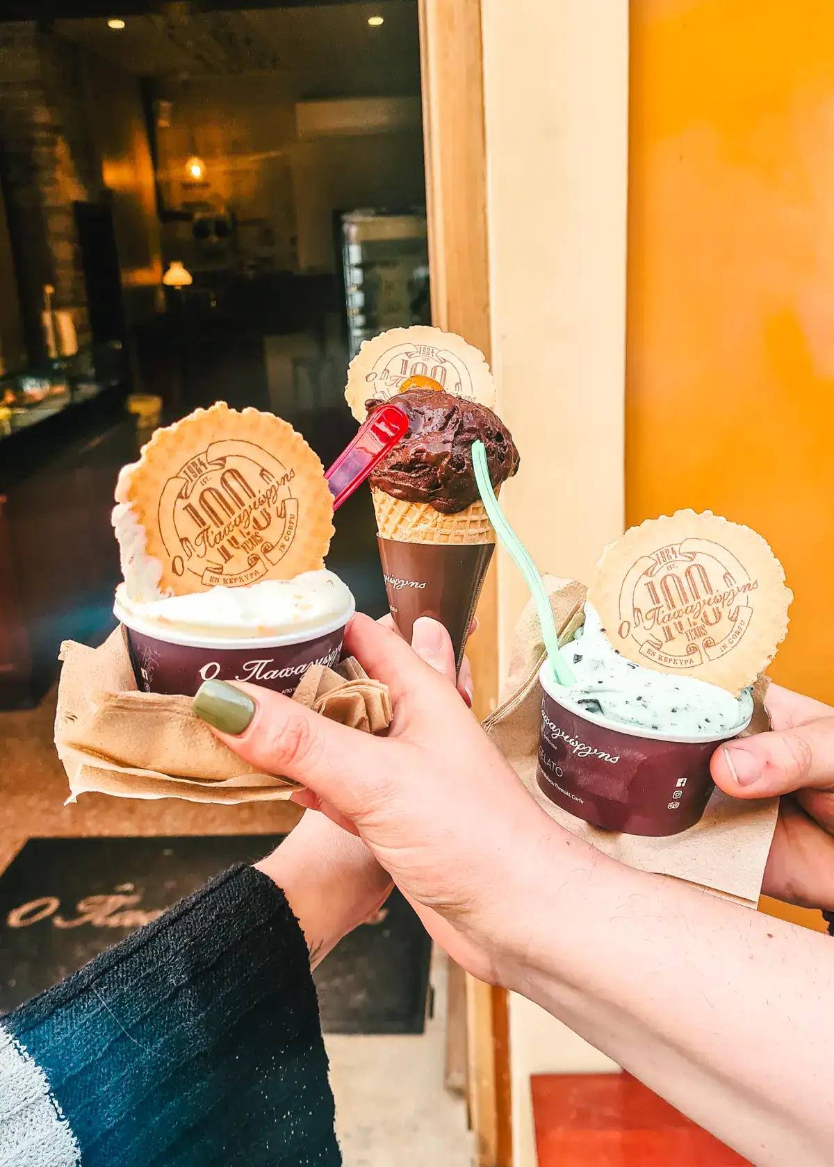 Three hands holding ice cream with a round twill with the shop logo and a spoon, in front of Papagiorgis Patiserie-Gelateria, the best ice cream shop in Corfu Old Town.