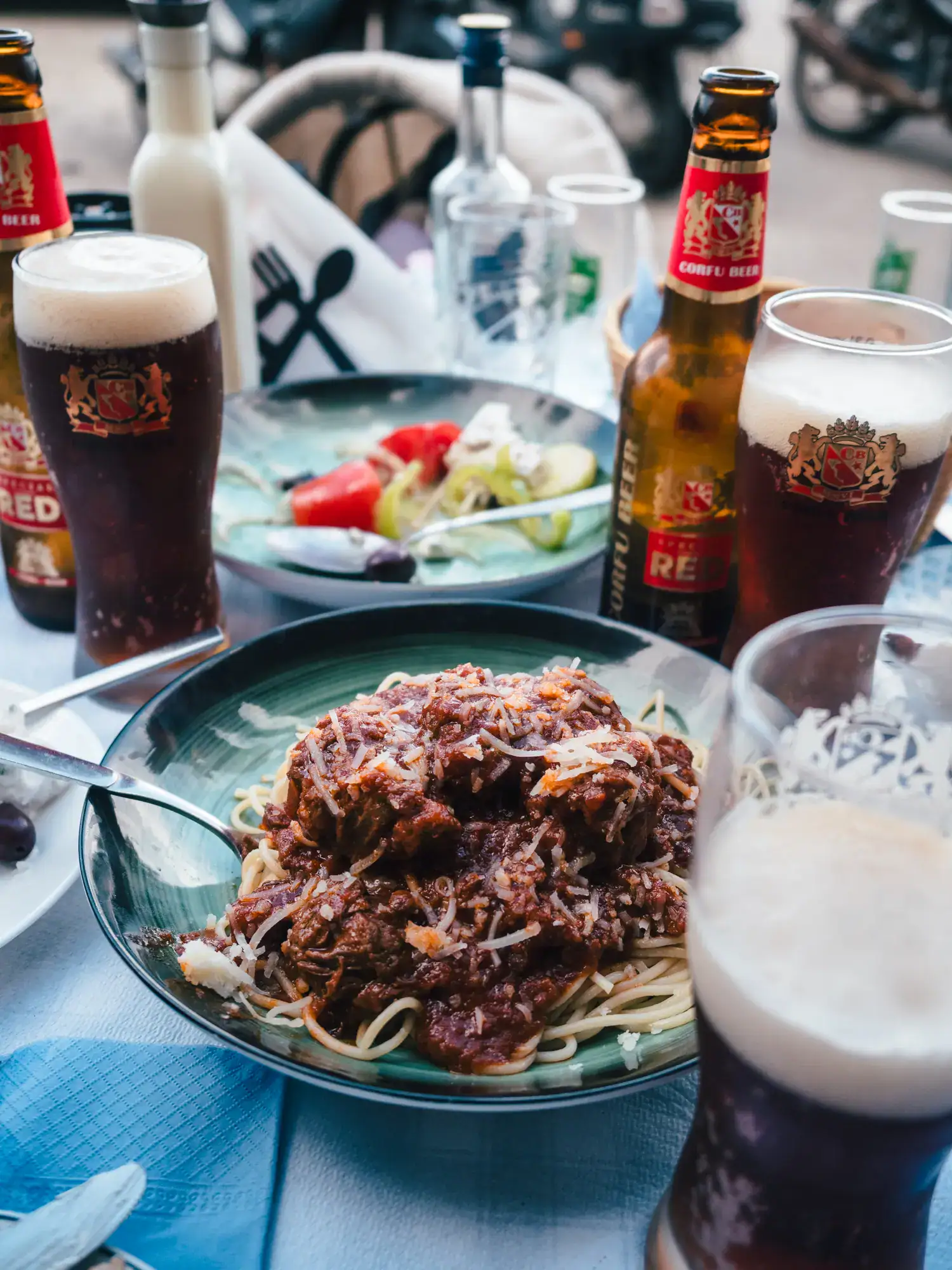 Close up of a green bowl of pasta and ragù, greek salad in the background, three glasses of Greek beer with foam, one flask with brown label on a table with blue table cloth, at Mouragia, one of the best restaurants in Corfu Old Town.