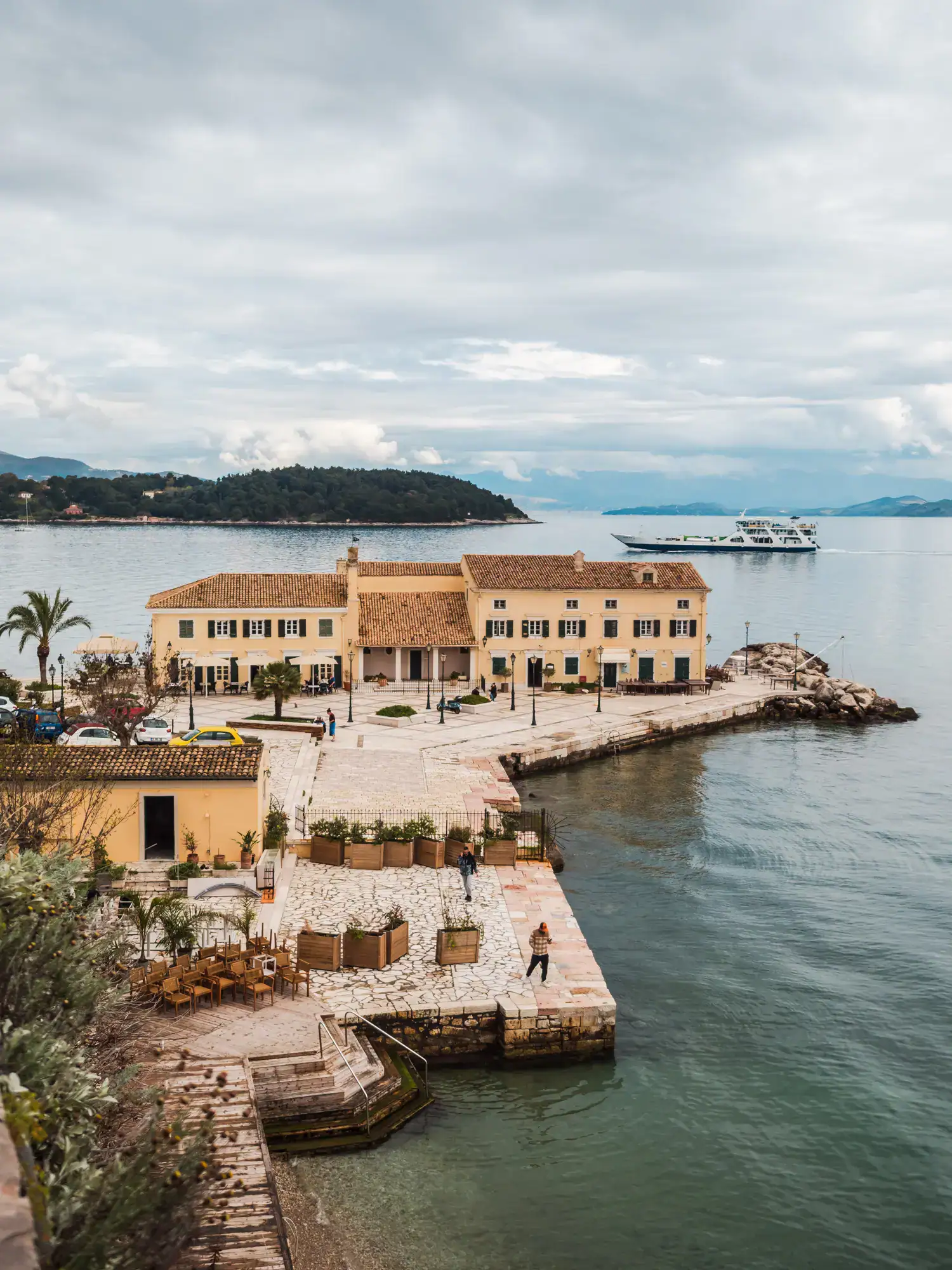 Looking down on a yellow building with a restaurant on a stone wharf, next two Faliraki Beach, with an island in the background and a ferry going by, during a day in Corfu Old Town.