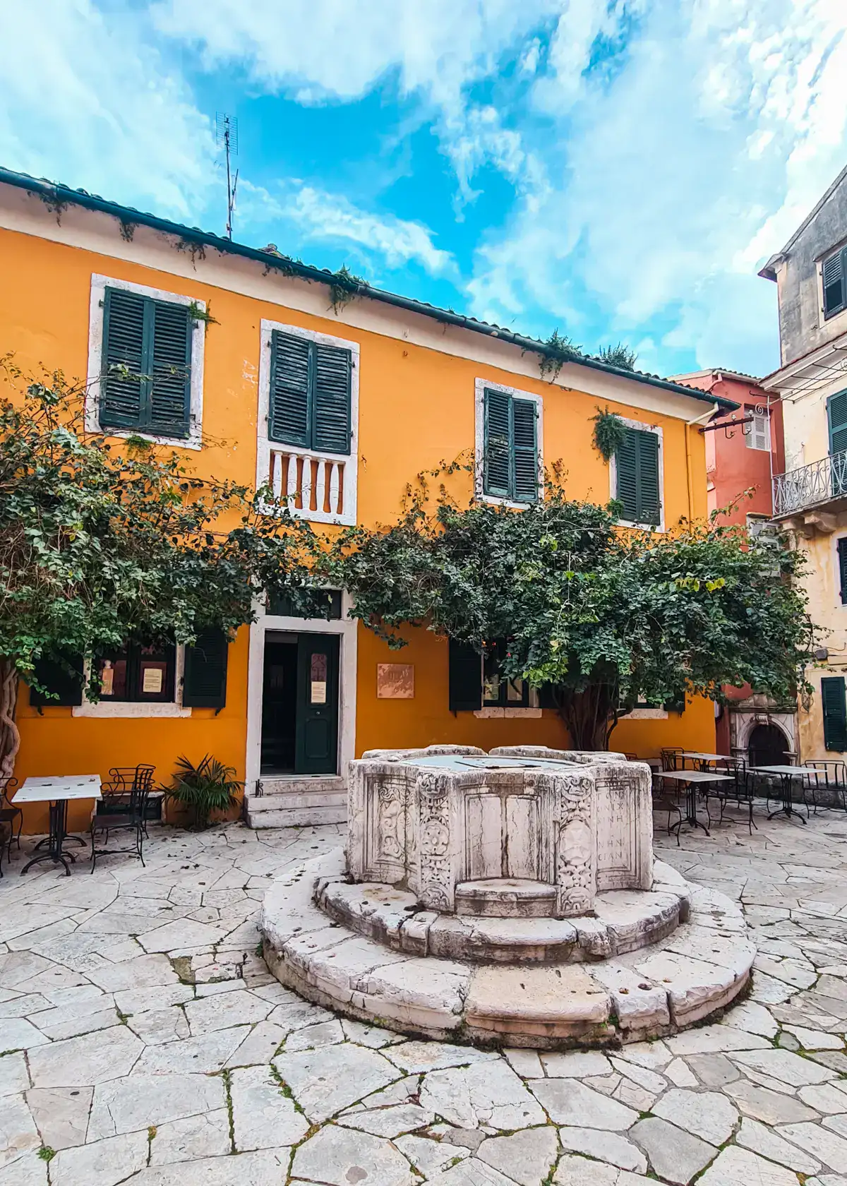 Old Venetian Well in Kremasti Square, with green tress and an orange building in the background, seen during the perfect Corfu Old Town Itinerary.