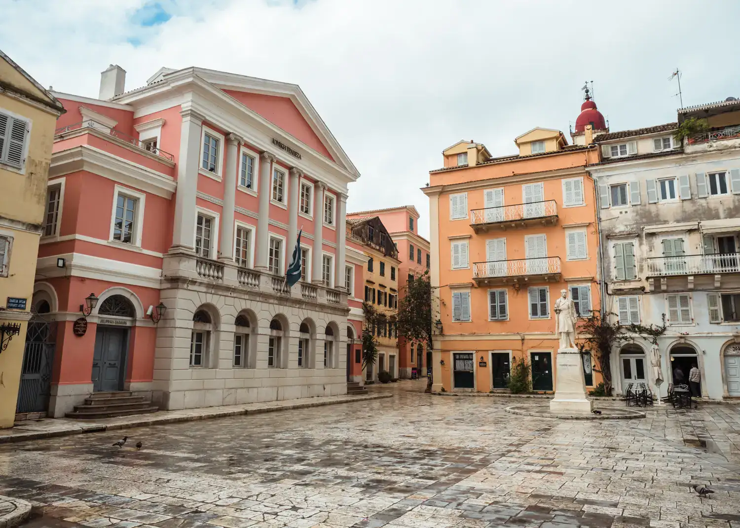 Tiles square with a white statue, a pink bank on the left and an orange building behind the statue, Plakada t' Agioú Spiridona Square in Corfu Old Town.