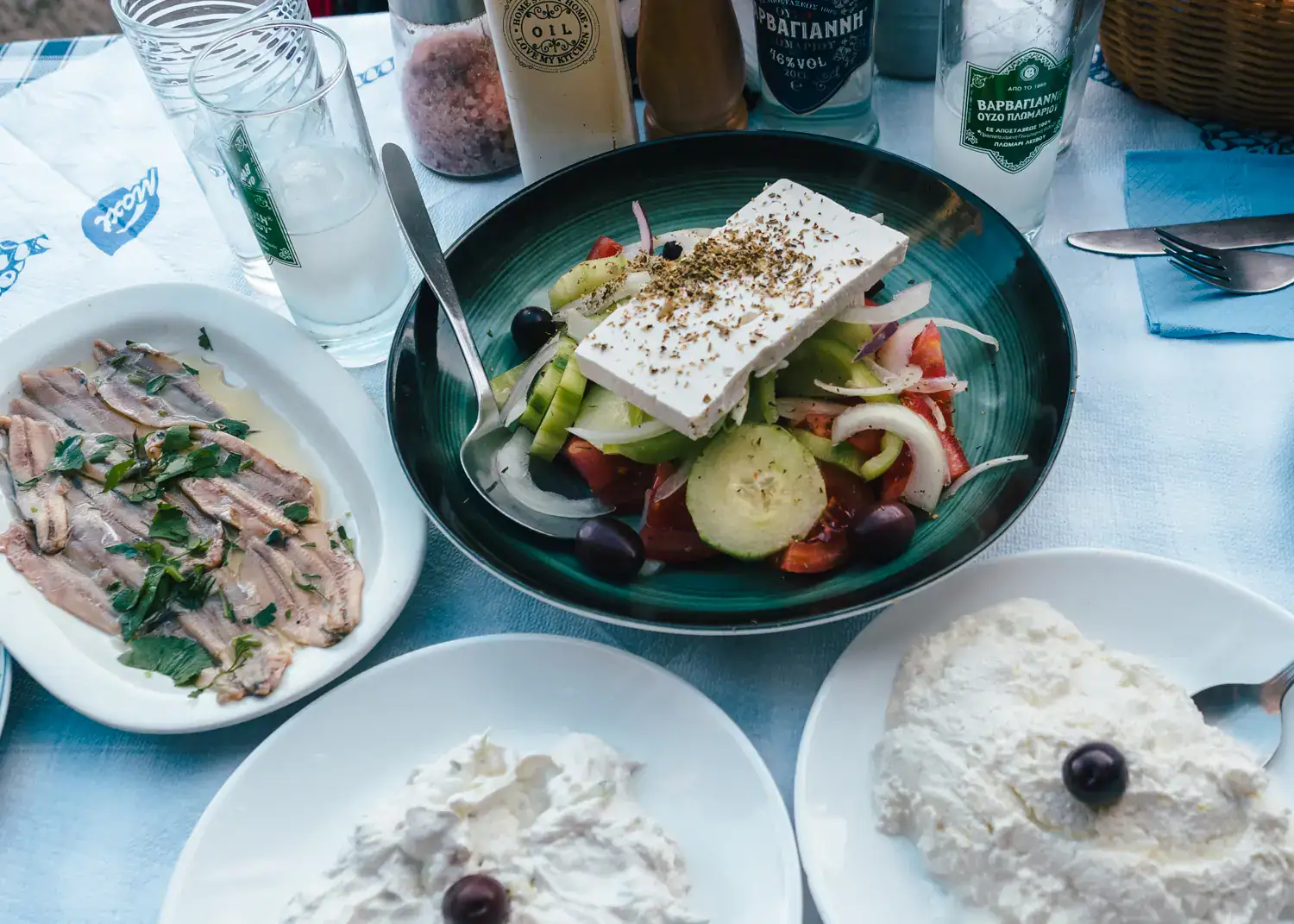 Close up of two white plates with white dips topped with a black olive, green bowl with Greek salad, white plate with anchovies, on a table with light blue tablecloth, at Mouragia, one of the best traditional restaurants in Corfu Town.