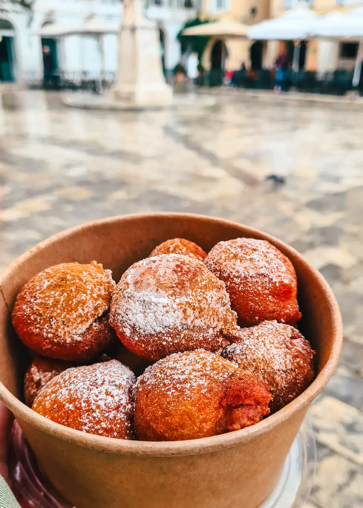 Close up of brown box with 8 Loukoumades, small Greek donuts, in a tiled square on a gloomy day in Corfu Old Town.
