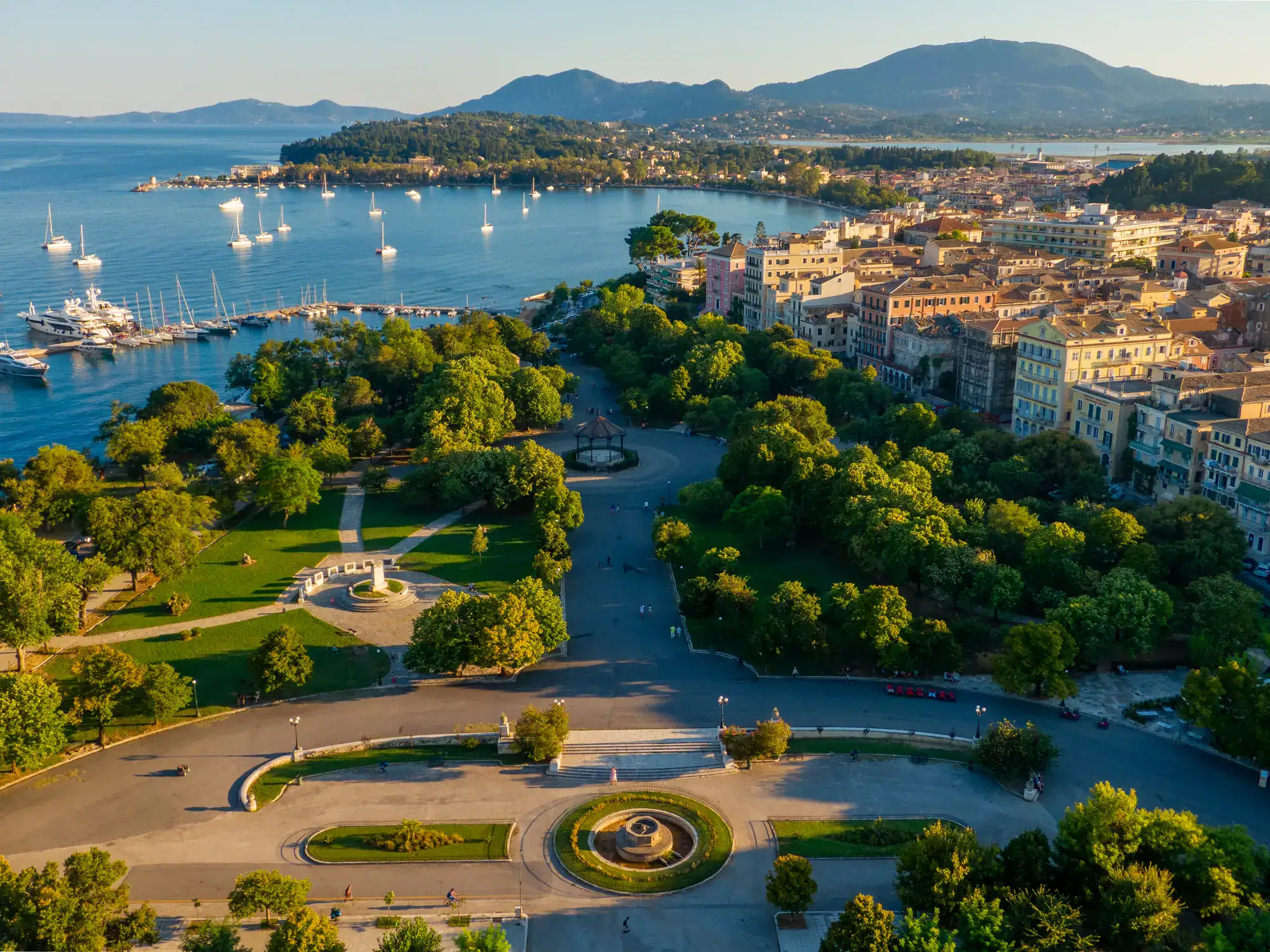 Aerial view of Spianada Square with a fountain and green trees, next to Corfu Old town, with the ocean and white boats in the background.