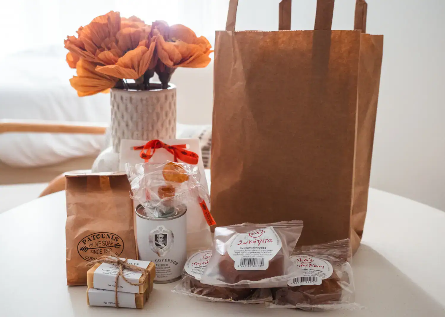White was with orange flowers, brown paper bag, olive oil soap, small white olive oil bottle, three date cakes on a white table, souvenirs bought during one day in Corfu.
