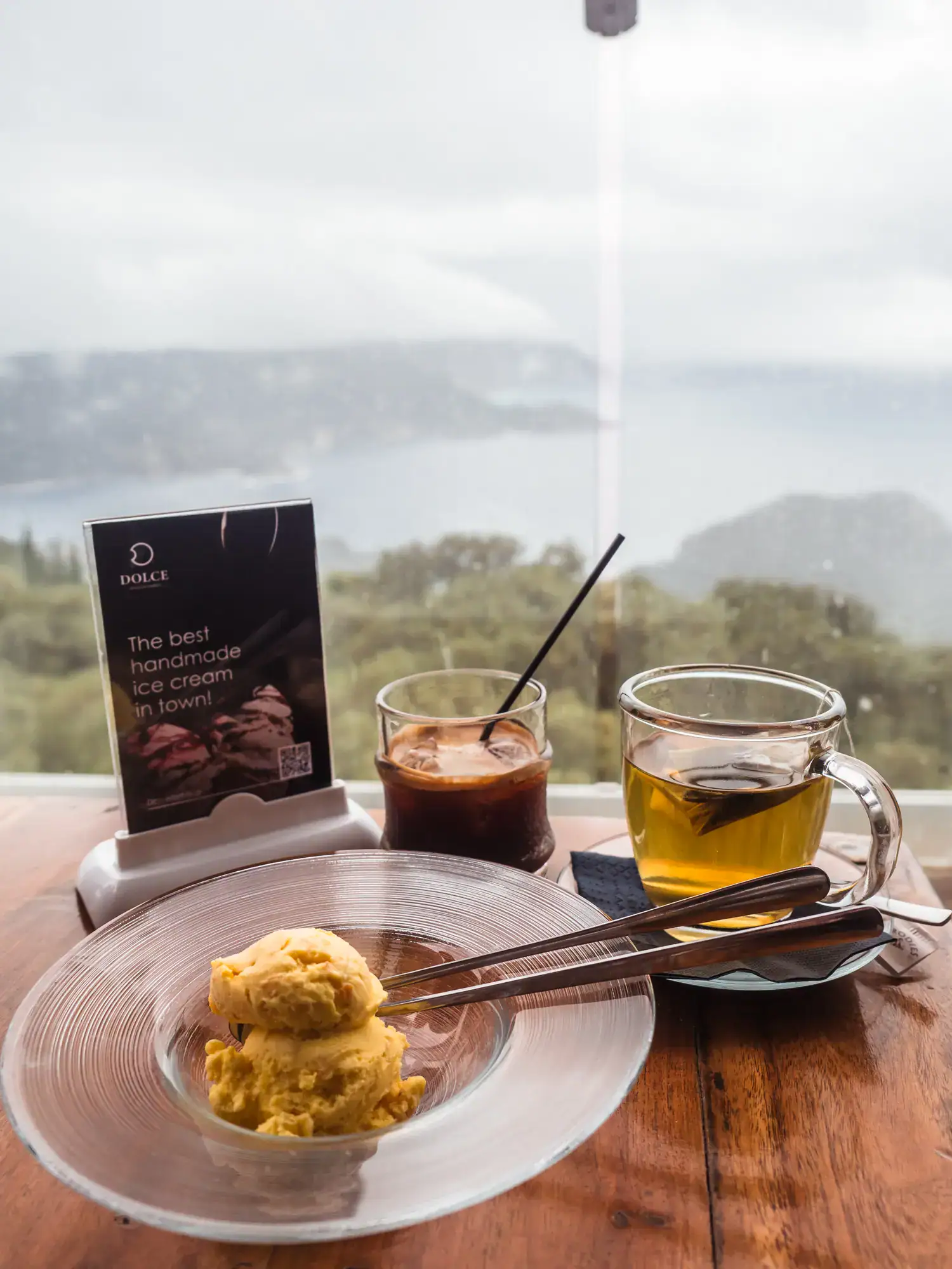 Yellow Kumquat ice cream in a clear bowl with two spoons, green tea and iced coffee on a table with the view of green forest and ocean in the background at Dolce Caf&eacute; in Lakones Village, during a one day Corfu itinerary.