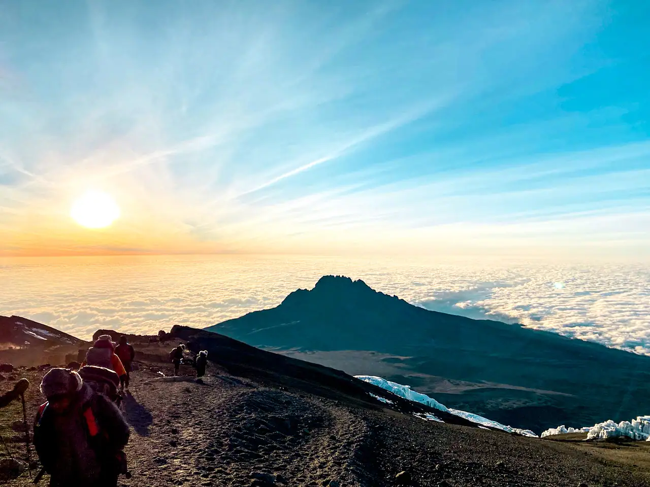 Path with people hiking up and down Mount Kilimanjaro during a yellow sunset on a blue sky, with a mountain peeking out from the white clouds in the background.