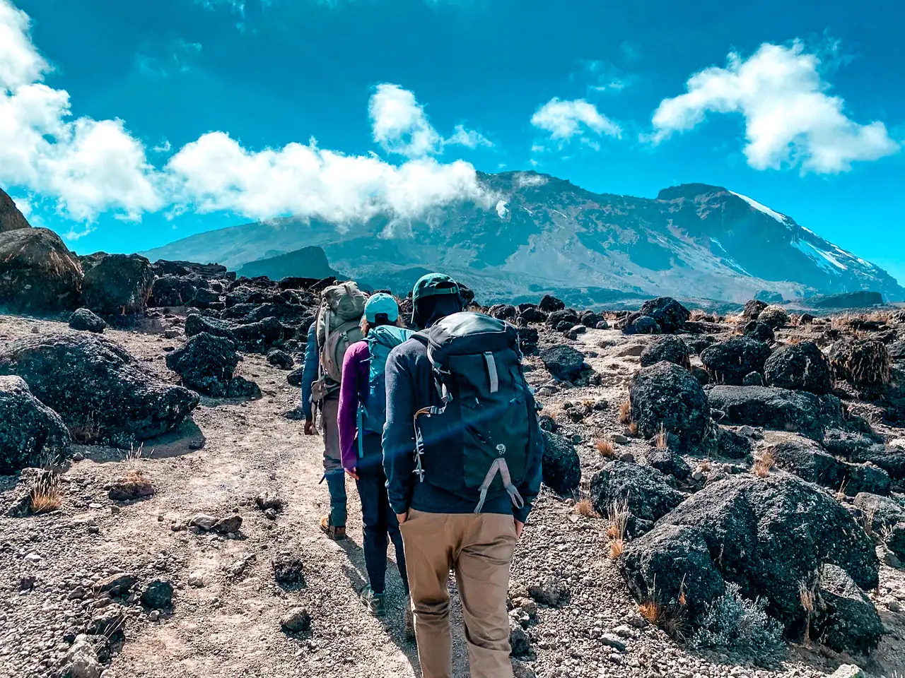 Three people with backpacks hiking up a dirt path with black volcanic rocks on both sides with Mount Kilimanjaro partly covered by white clouds in the background.