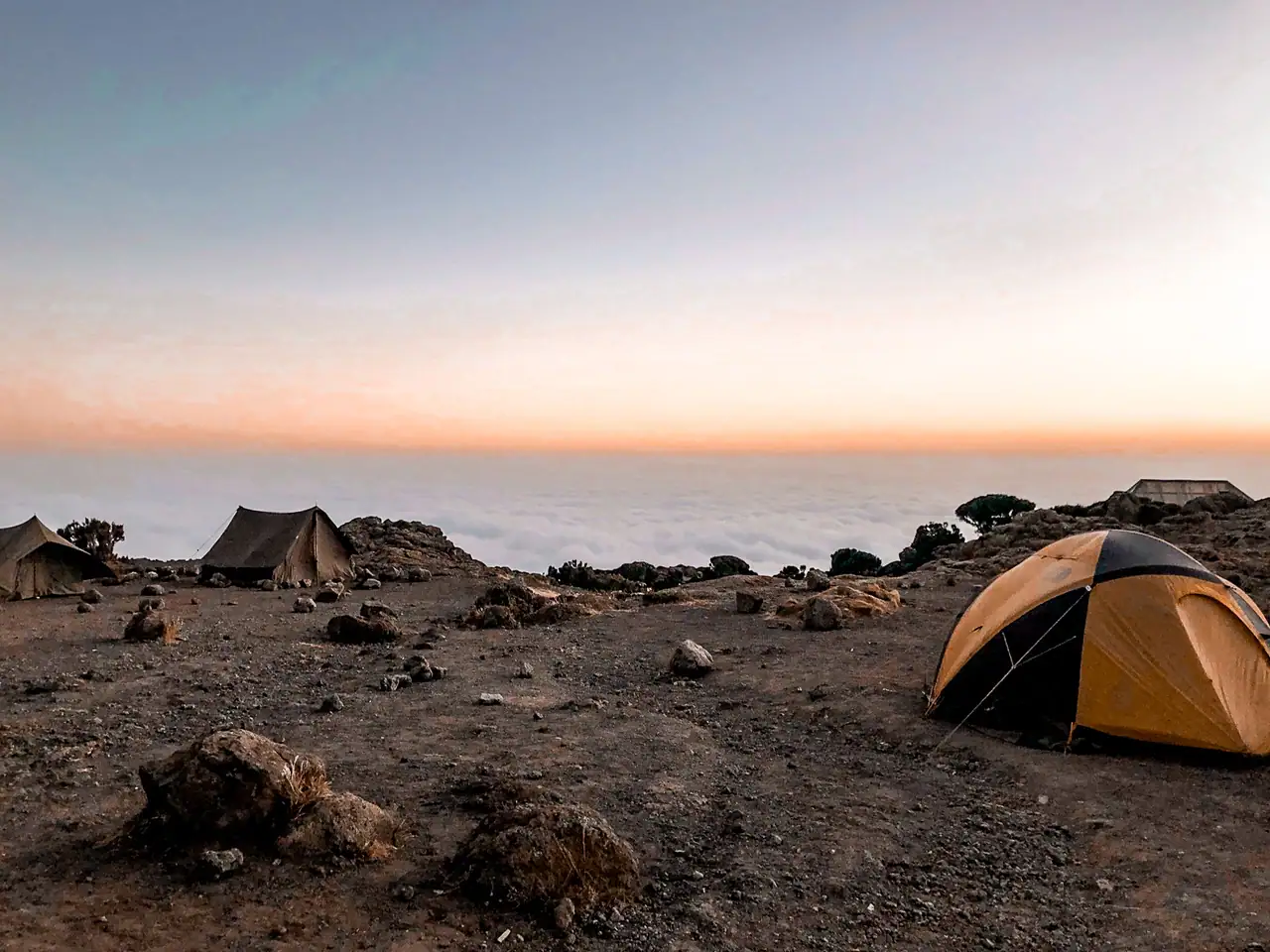One orange and black tent and two brown tents in the background on a dirt campsite over the clouds at sunset. Hiking Kilimanjaro, what you need to know.