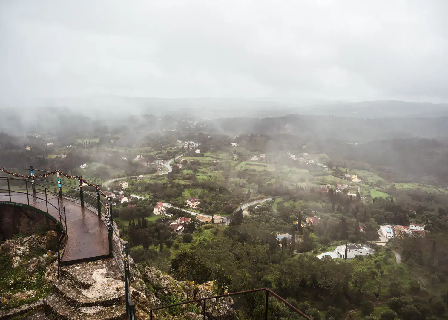 Misty view of the lush green countryside in Corfu on a rainy day from the rounded Kaiser's Throne Viewpoint with metal railing, during a one day Corfu Itinerary.