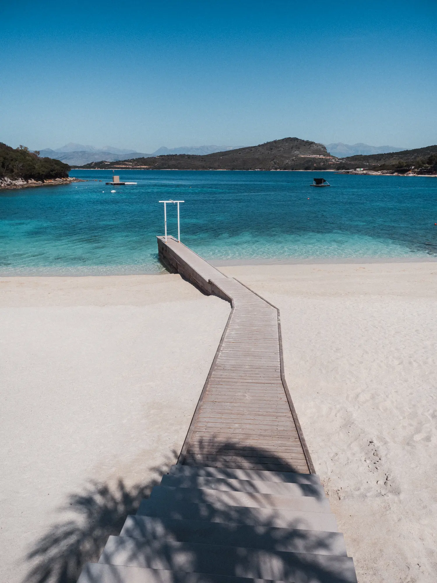 Thin wooden jetty going across the white, clean sand into the blue ocean with a plan tree shadow on a sunny day at Ksamil Beach.