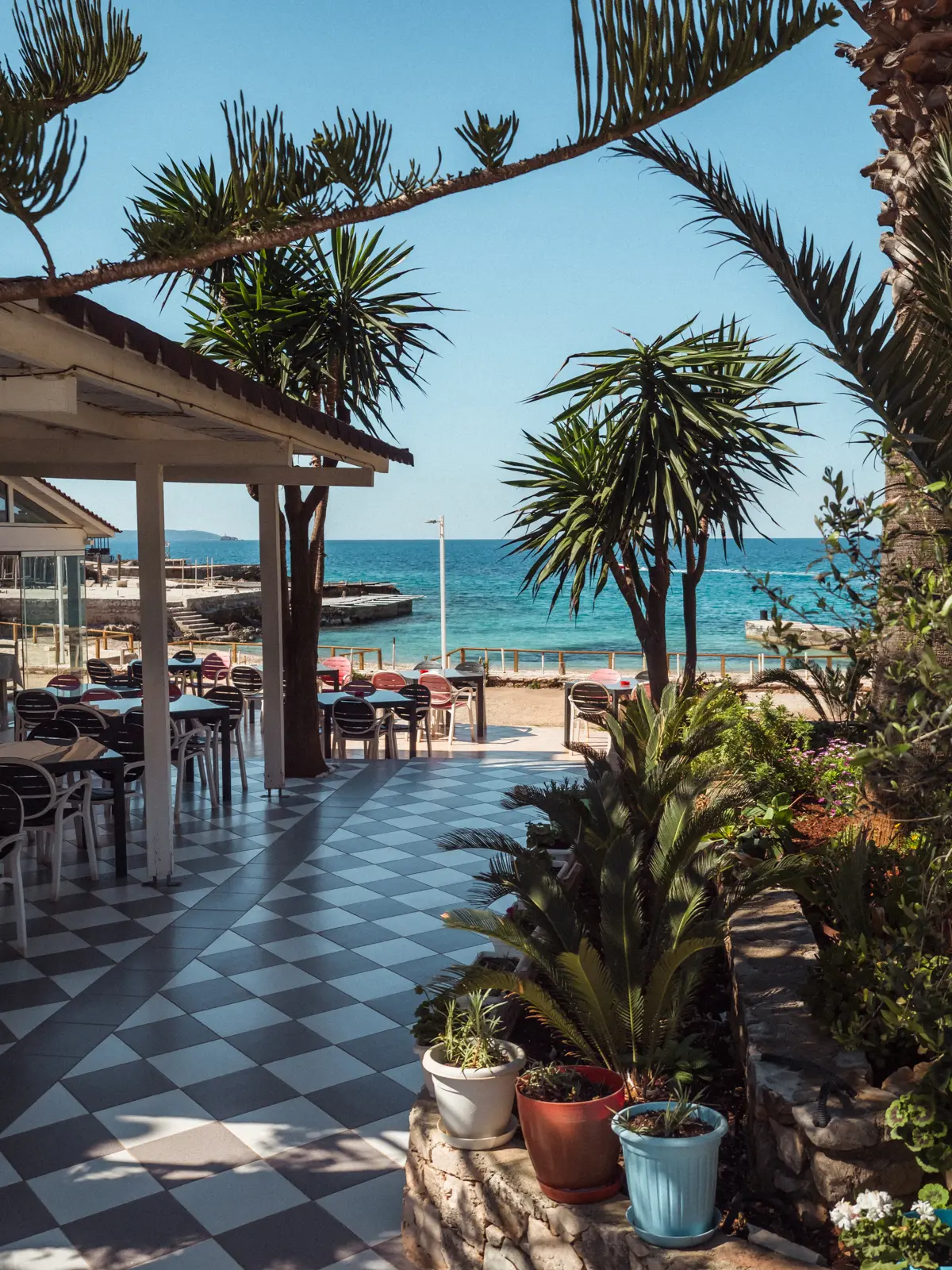 Restaurant by the beach with grey and white checkered floor with palms and green plants on the right and the turquoise ocean in the background on a sunny day in Ksamil.