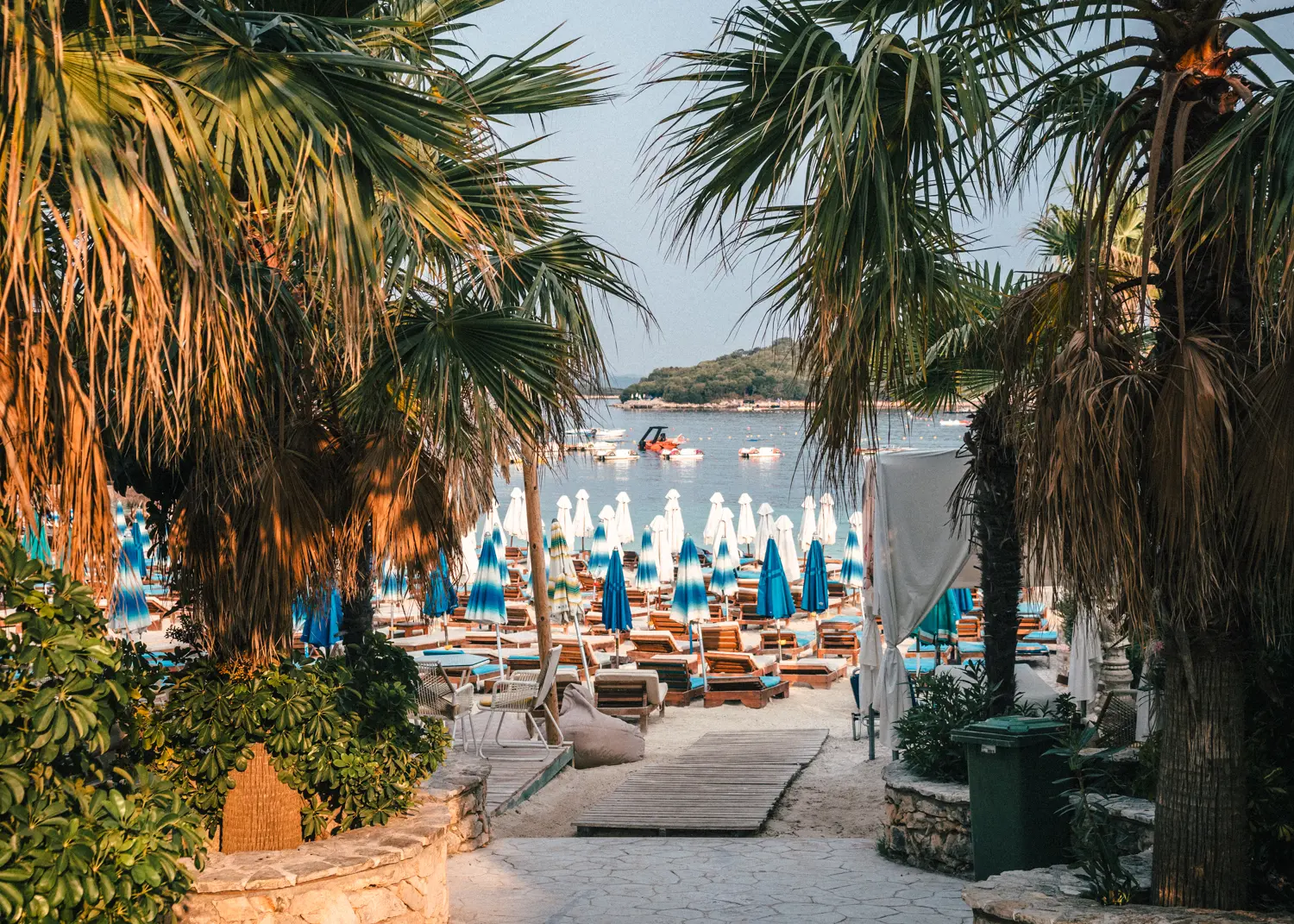 Looking through palm trees on a beach with wooden subbed and blue and white parasols with the ocean and an island in the background, Ksamil Beach guide.