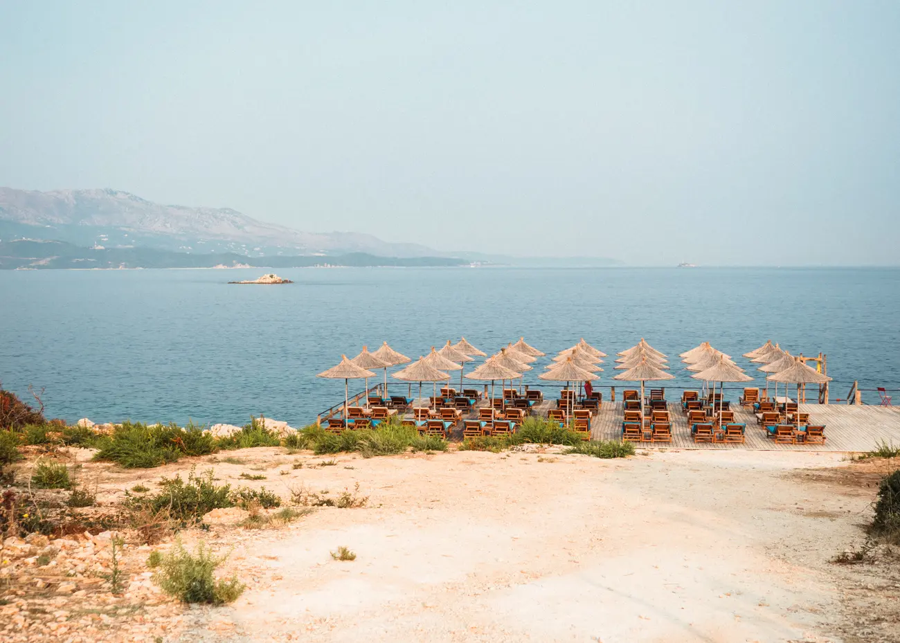 Square area of wooden sunbeds and straw umbrellas along the rocky coast in Ksamil, Albania.