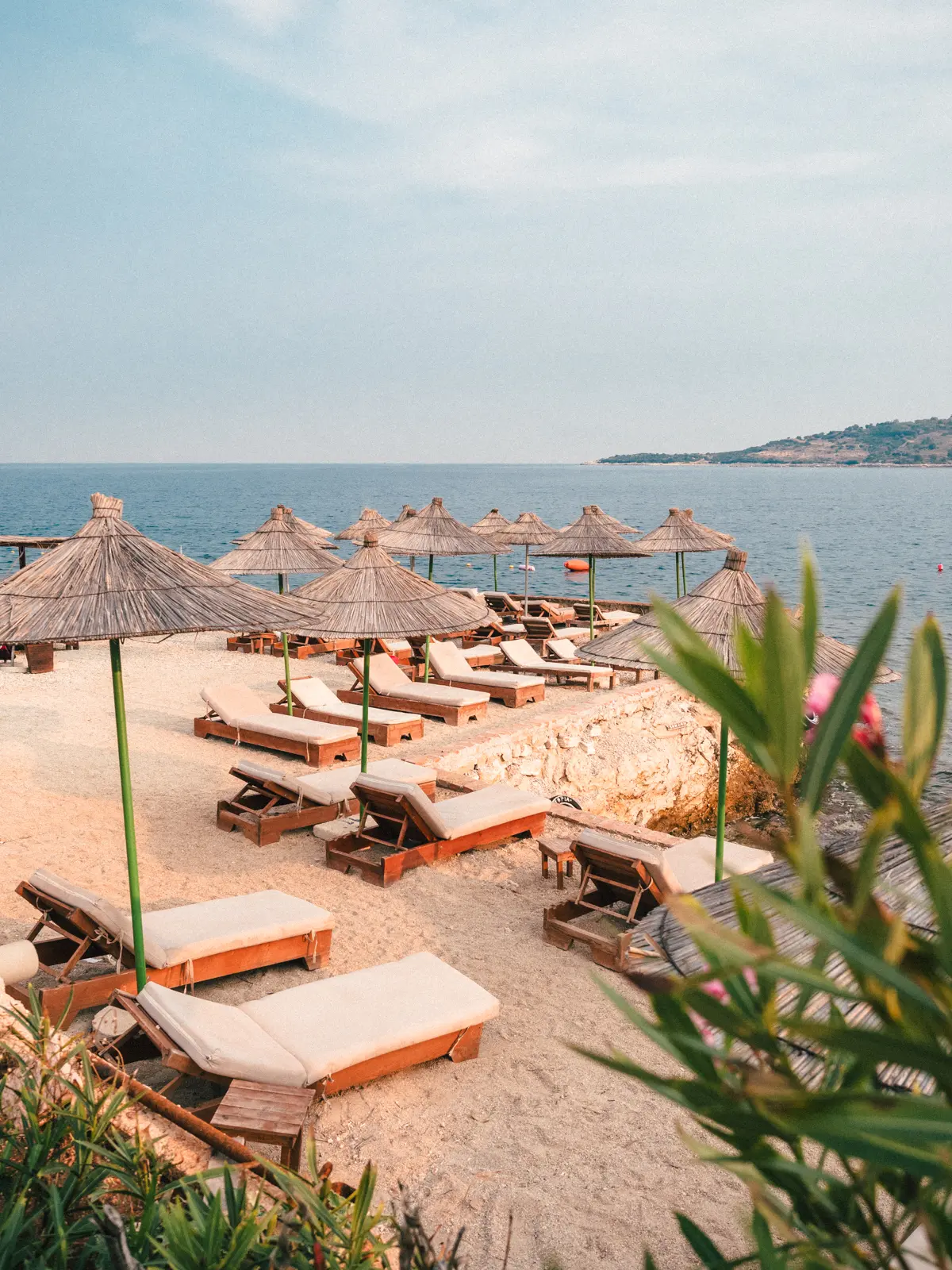 Wooden sunbeds with white pillows and straw umbrellas on a rock area with sand on top in Ksamil.