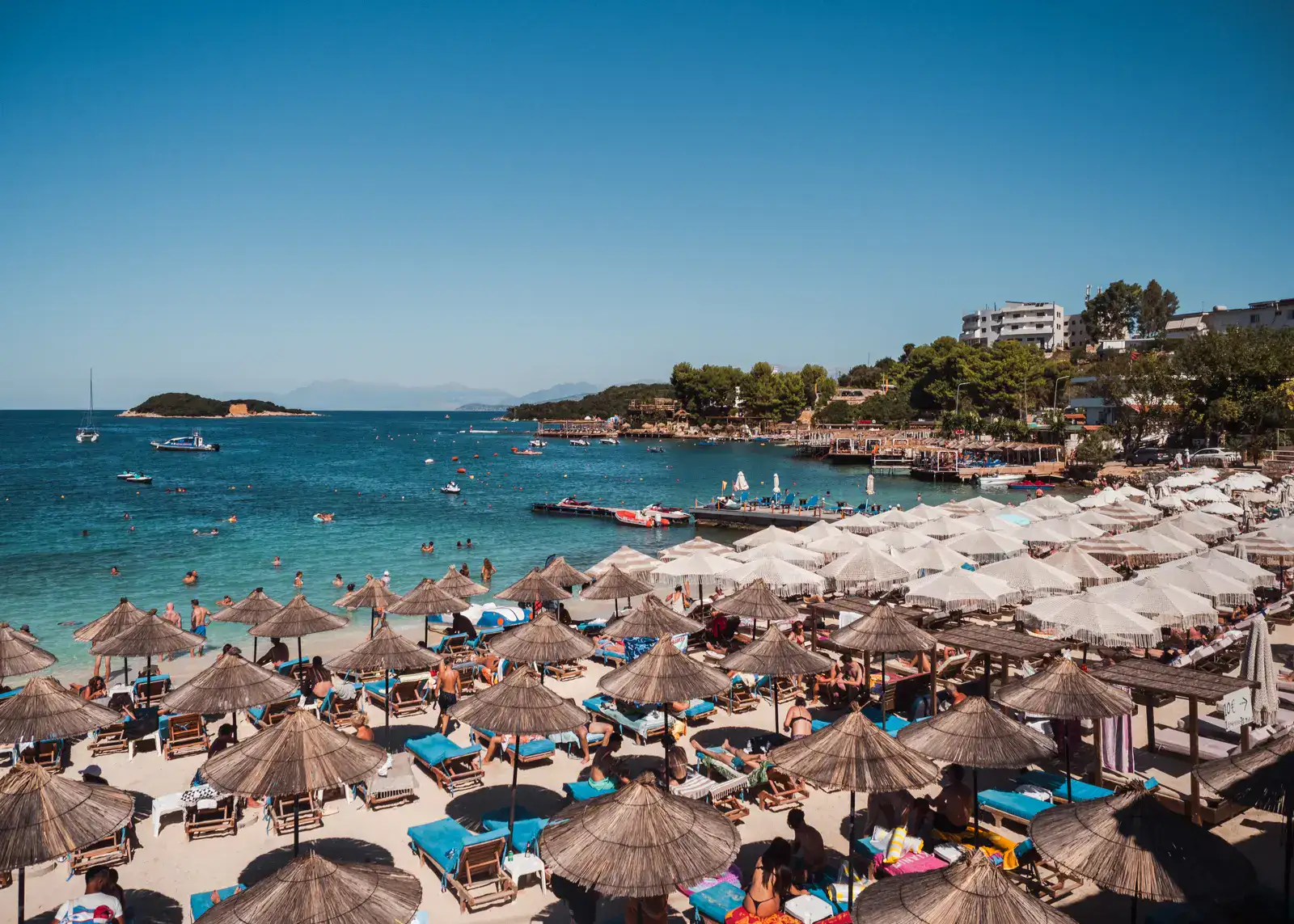 View of the busy Castle Beach in Ksamil, covered in half straw and half white parasols, on a sunny day with blue skies.