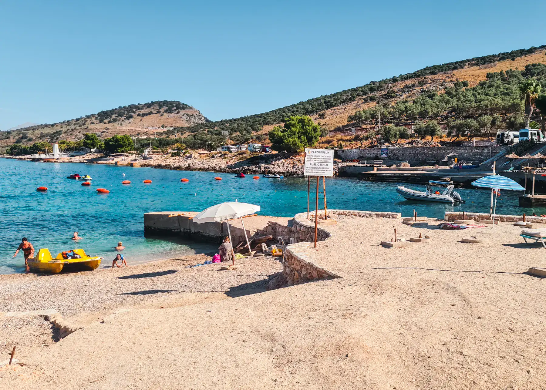 View of public beach in Ksamil, built up area with sand next to a small pebbled beach, yellow paddle boat, a few people in the water, hills with some greenery in the background.