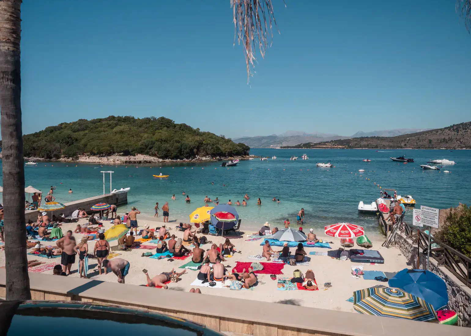 White sand beach covered in people on towels, some parasols, turquoise water and a small, green islet in the background. Ksamil public beach.
