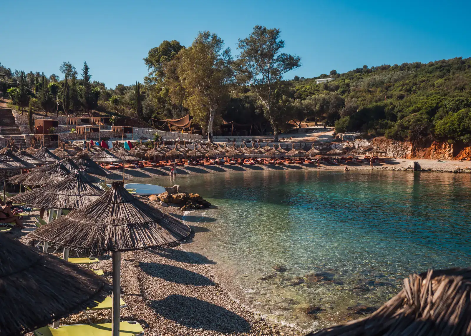 Early morning view of the half-moon shaped Last Bay in Ksamil, with orange bean bags and straw parasols, clear bluish green water on a sunny day.