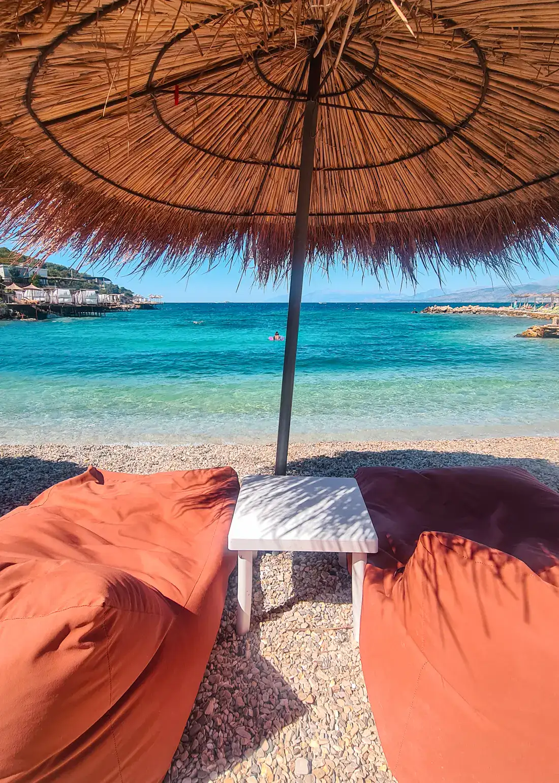 Two orange bean bags with a white table in the middle, under a straw parasol, looking our at the clear turquoise water at Augustus Beach in Ksamil.