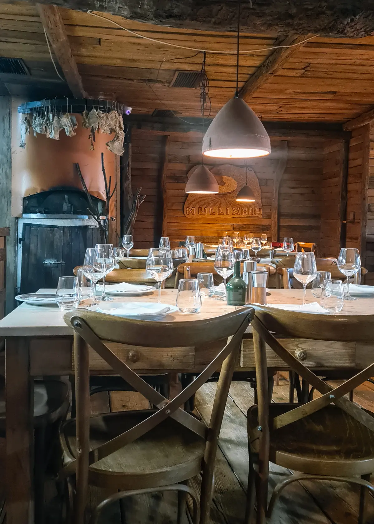 Four wooden chairs around an old wooden table in an old wooden house with an oven in the background and several plates and wine glasses on the table at Mullixhiu Restaurant in Tirana, Albania.