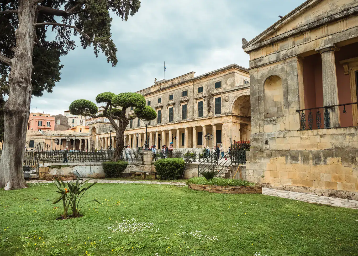 View of Museum of Asian Art, set in a sandstone collared Neoclassical Palace with pillars, green grass and Japanese looking tree in front, in Corfu Old Town.