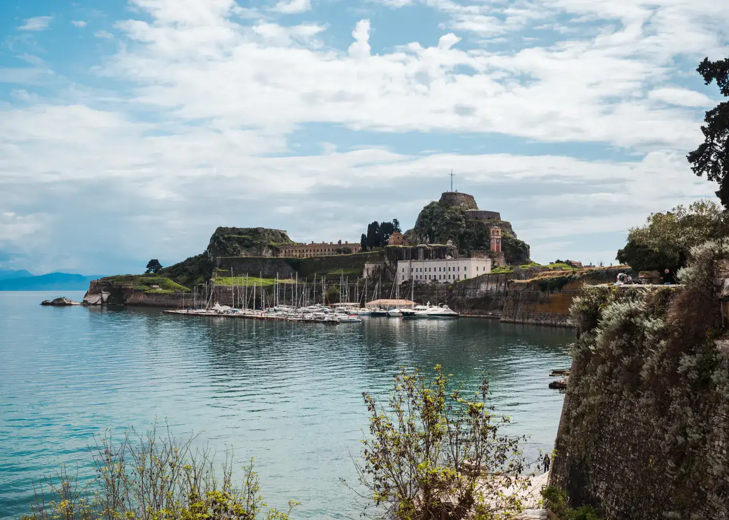 View of the Old Fortress on a headland in Corfu Town, with a cross on top, white sailboats next to it, seen during a one day Corfu itinerary.