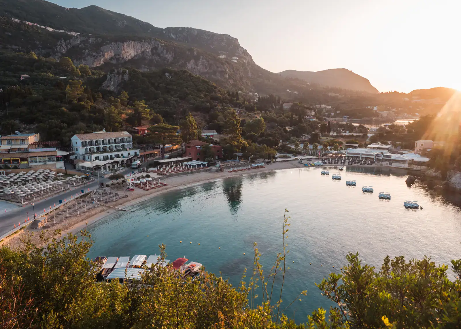 Early morning view from above of the half-moon shaped Paleokastritsa Beach with areas of sunbeds, calm blue water with reflections, hotels and restaurants along the back, and mountains in the background, sun shining on the right.