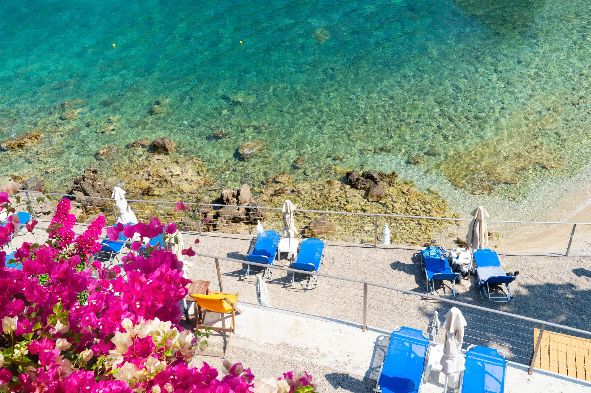 Blue sunbeds on a concrete patio by the shallow turquoise ocean at Paleokastritsa Beach in Corfu, dark pink Bougainvillea in the foreground, during a one day tour of the island.
