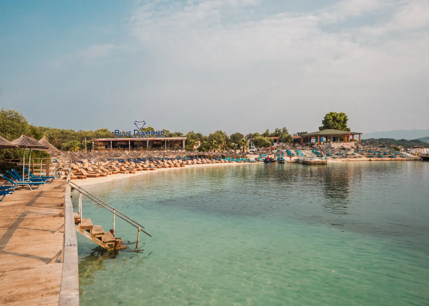 View of Puerto Rico Beach in Ksamil, with with a wooden deck on the left, light greenish turquoise water and a white beach covered in sunbeds with the Blue Diamond Beach Bar in the background.